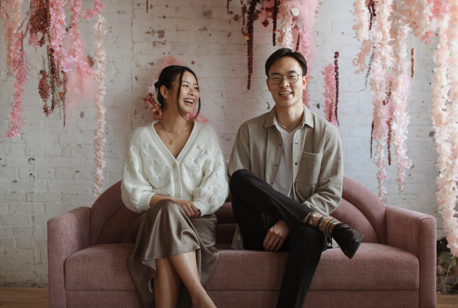 Couple hugs while sitting. Background white brick with pink flowers