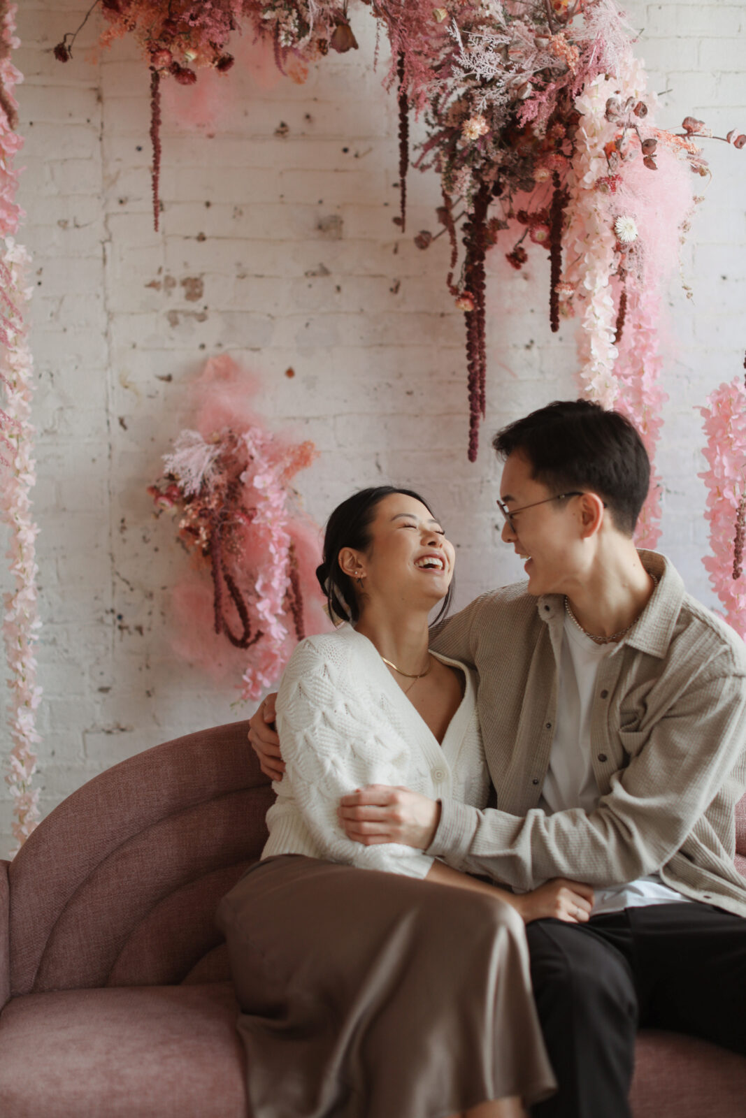 Couple laughs while sitting. Background white brick with pink flowers