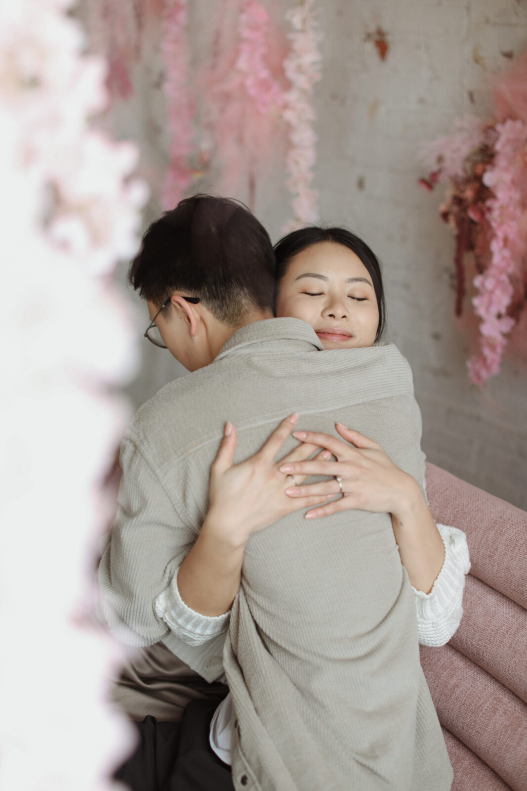 Couple hugs while sitting. Background white brick with pink flowers