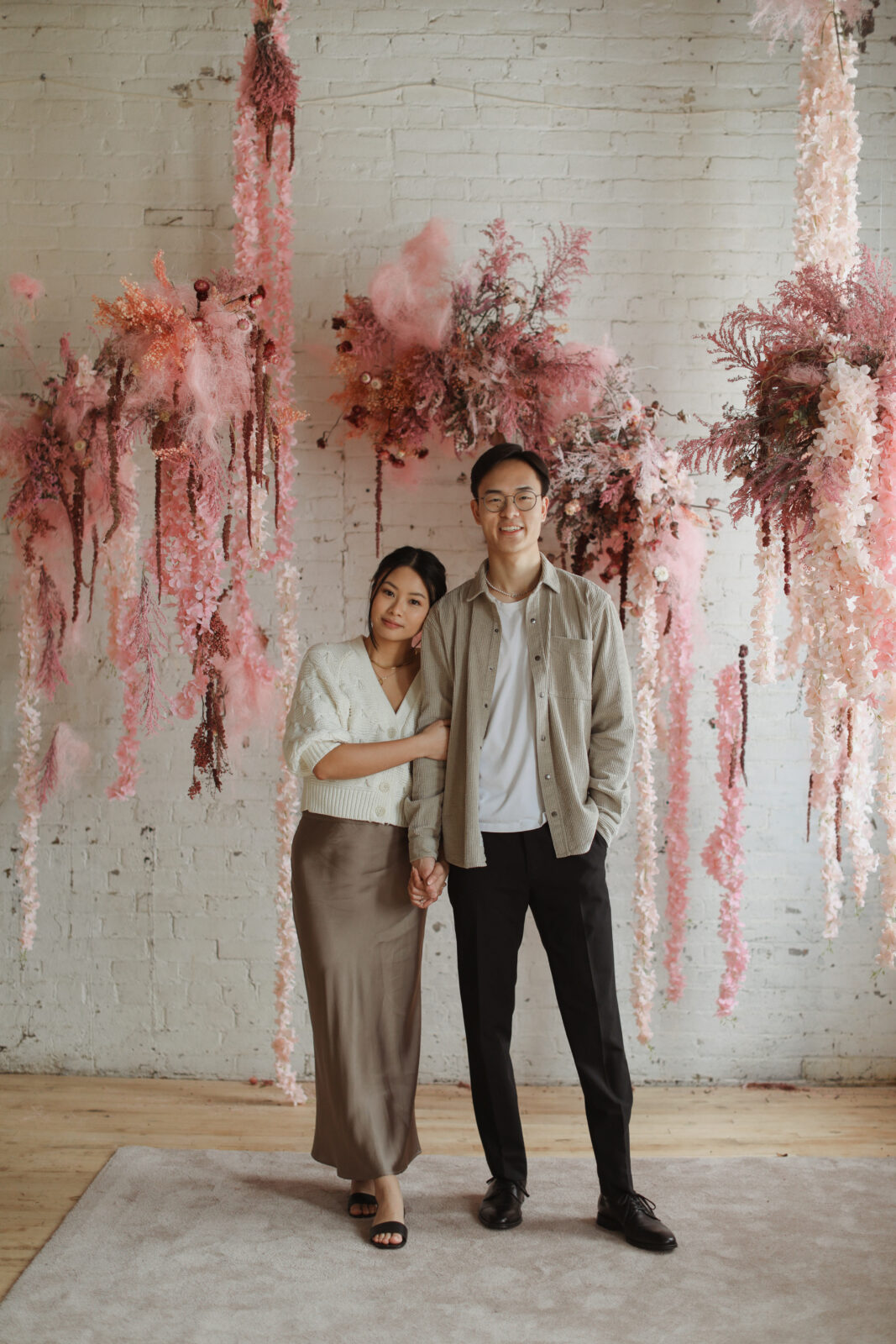 Couple smiling at camera. Background white brick with pink flowers