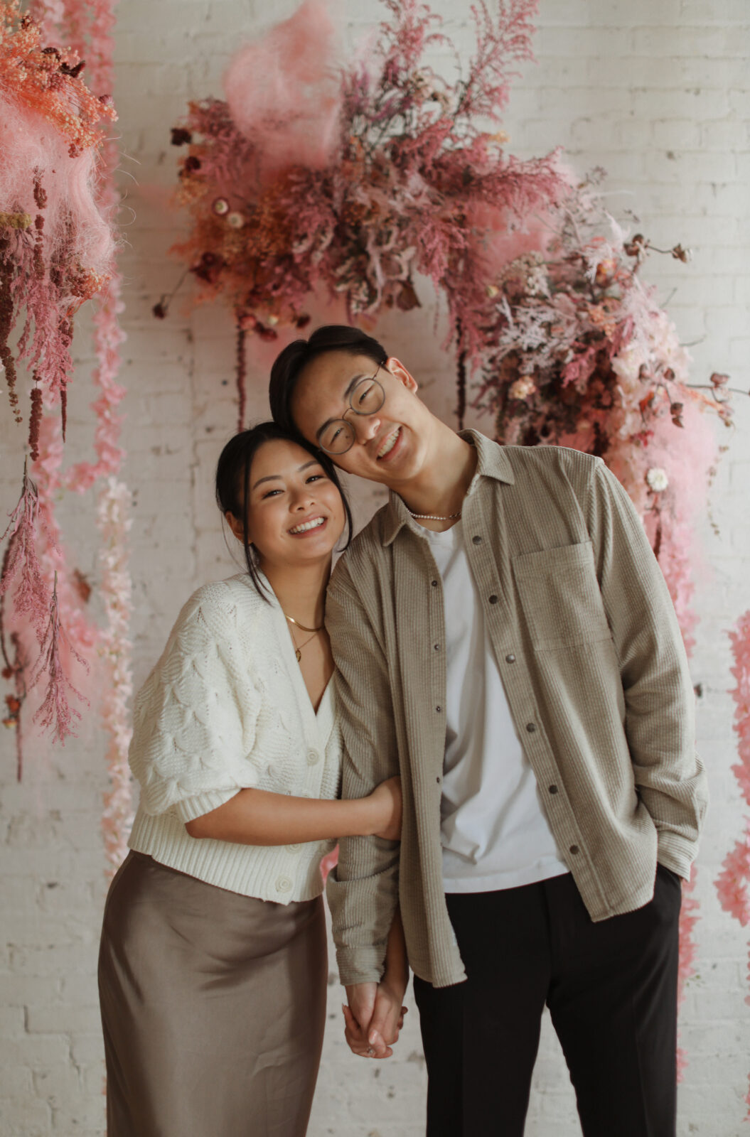 Couple smiling at camera. Background white brick with pink flowers