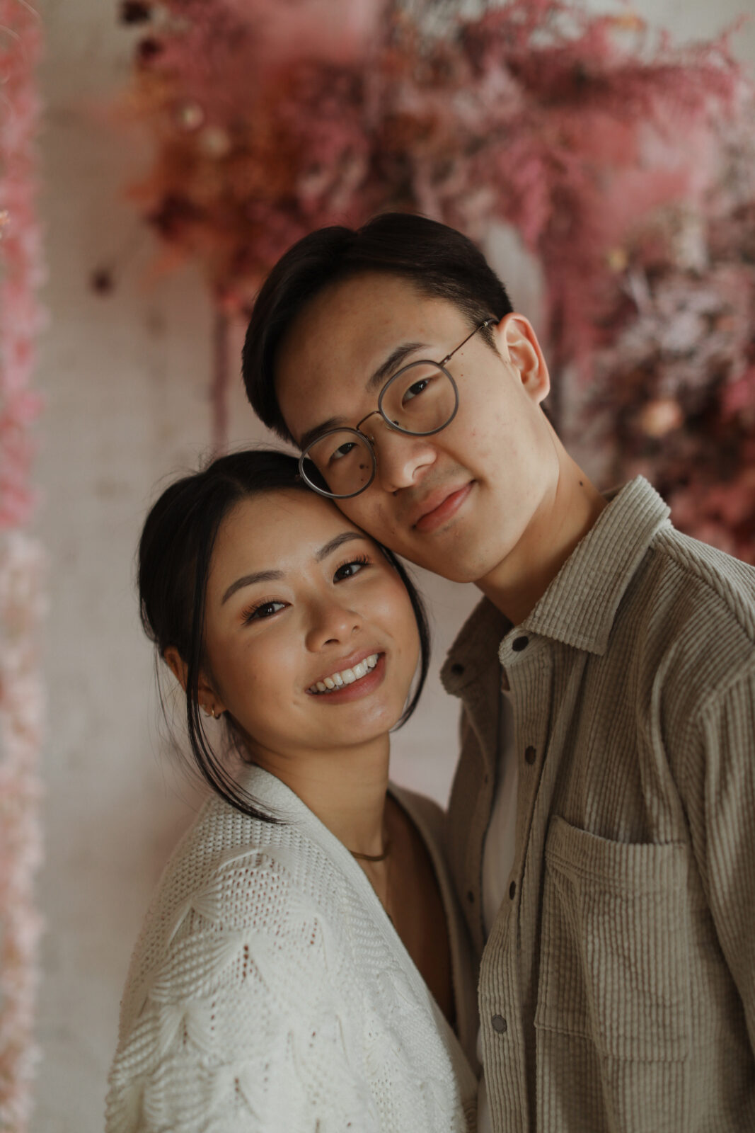 Couple smiling at camera. Background white brick with pink flowers