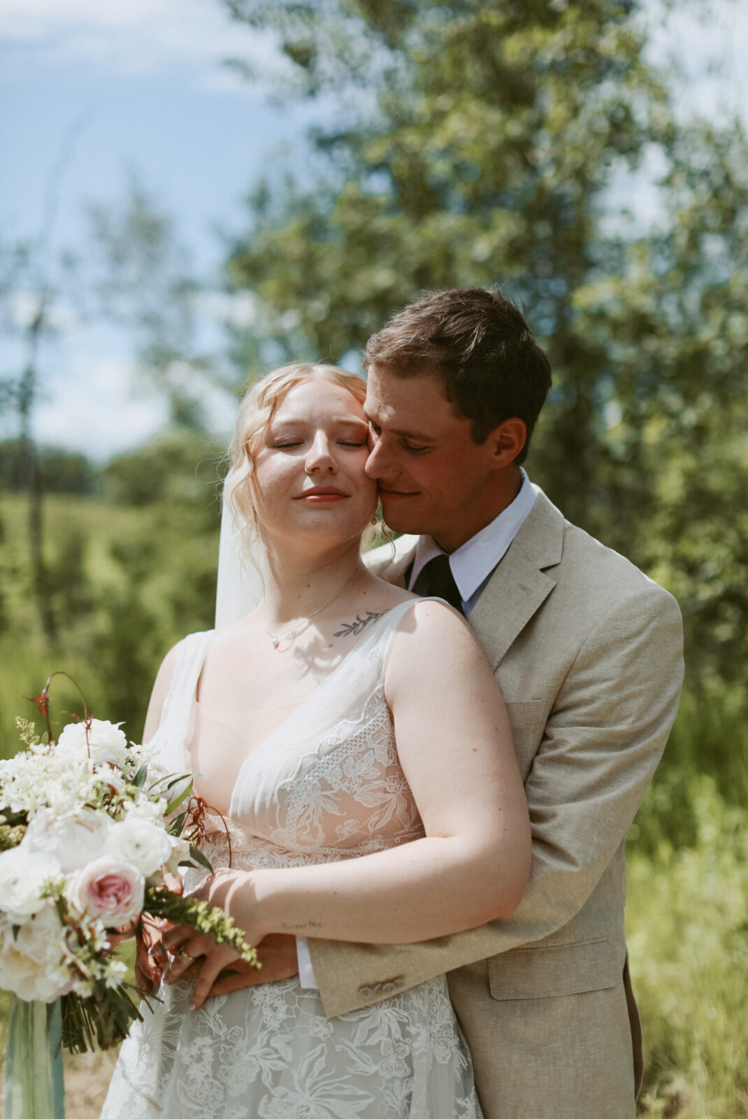 Groom holds bride around the waist. They both have eyes closed while sun shines on their faces. 