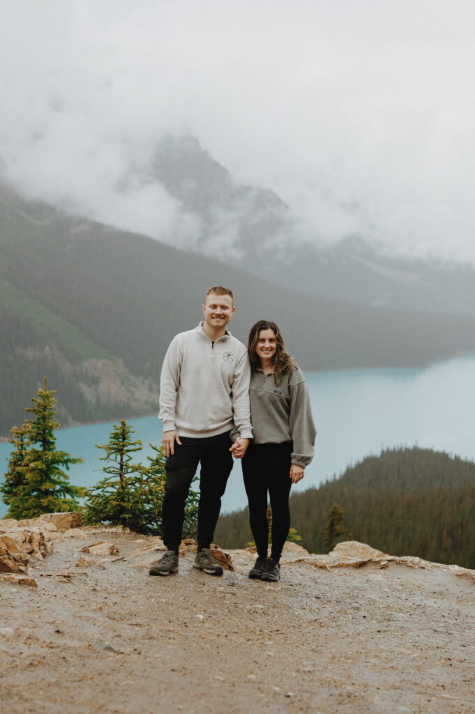 Man and woman hold hands on top of a mountain with a lake in the background. 