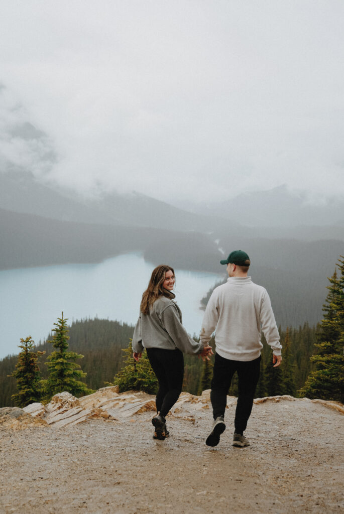 Man and woman hold hands on top of a mountain with a lake in the background. 