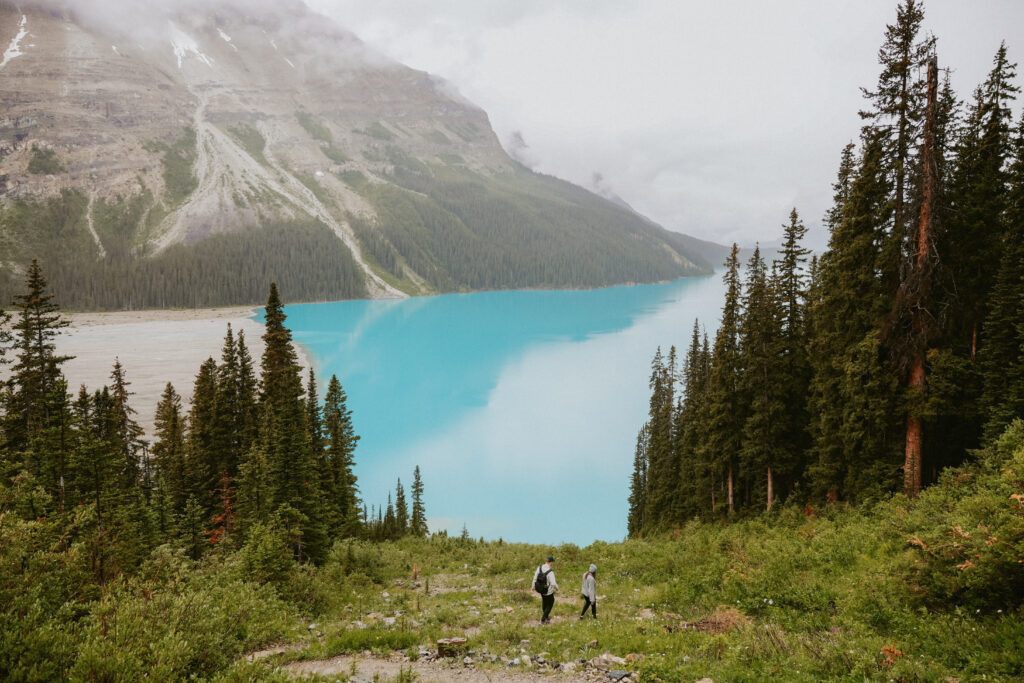 Couple hikes down mountain towards crystal blue lake. 
