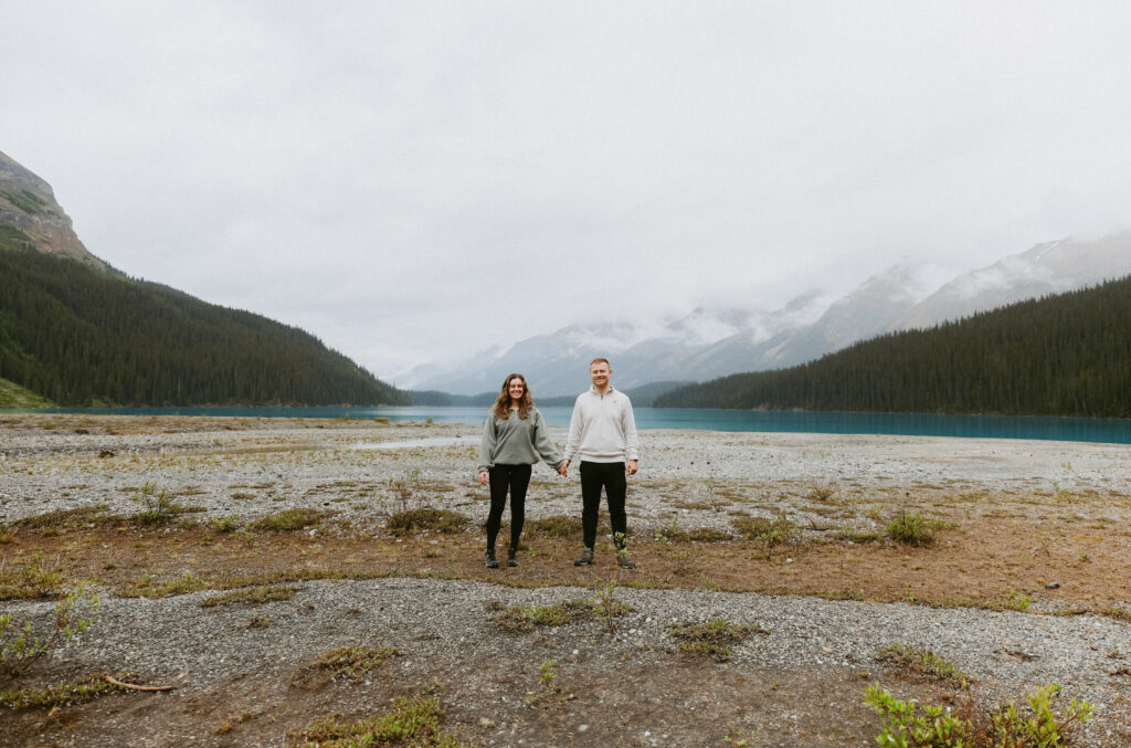 Couple holds hands on a rock shoreline in Alberta. Crystal blue lake in the background.