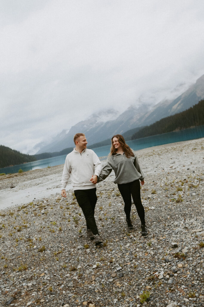 Couple holds hands on a rock shoreline in Alberta. Angle of image is askew with crystal blue lake in the background. 