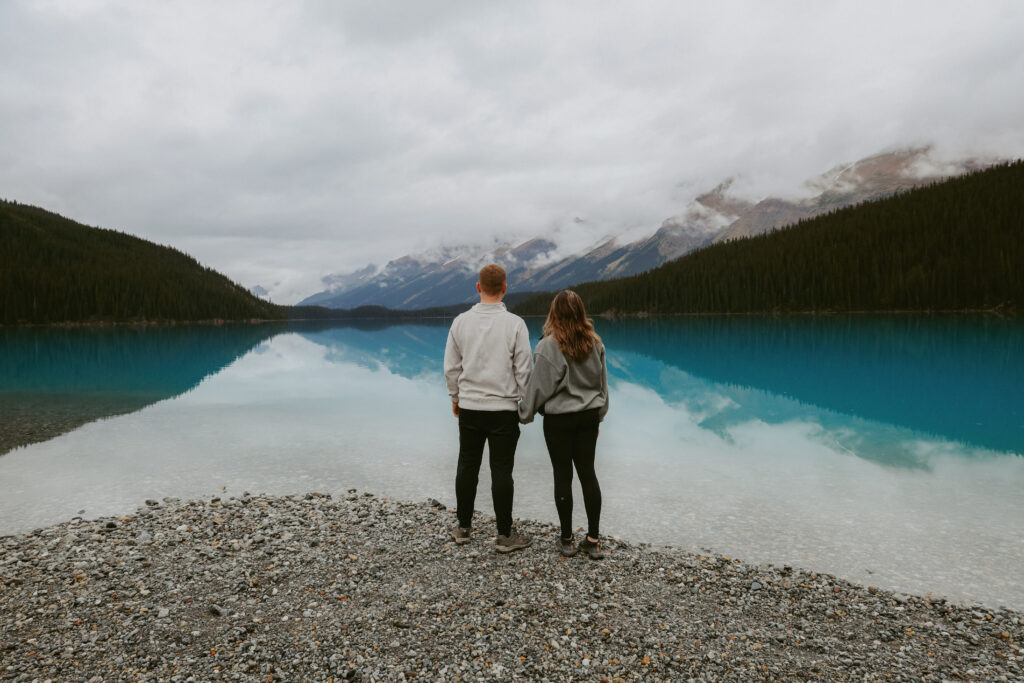 Couple holds hands on a rock shoreline in Alberta. Couple is standing with their backs to the camera looking at the lake. 