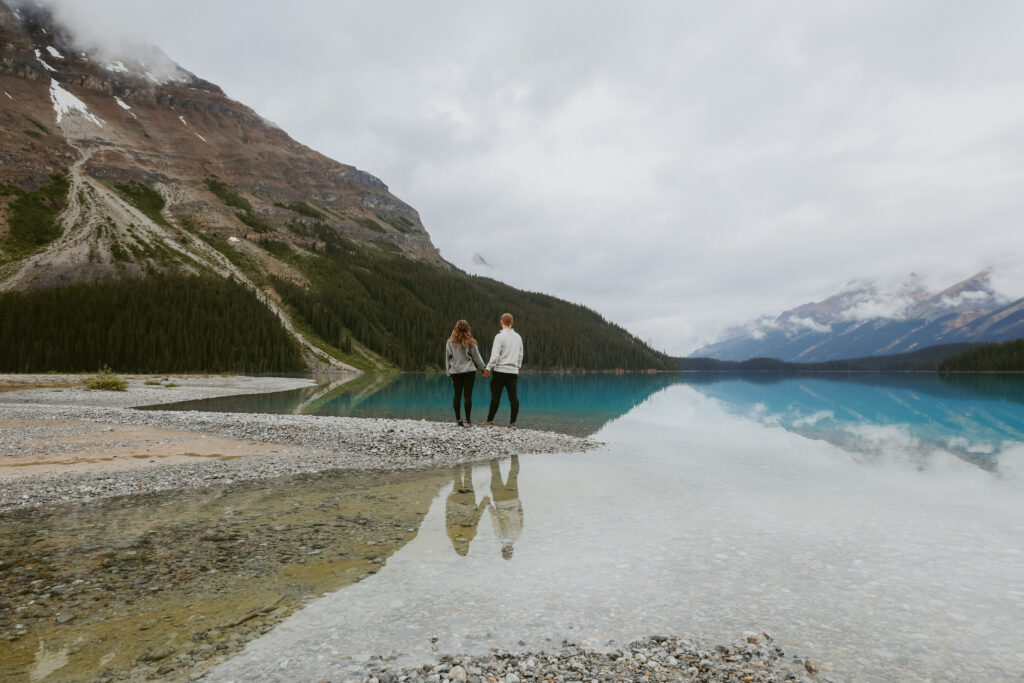 Couple holds hands on a rock shoreline in Alberta. Couple is standing with their backs to the camera looking at the lake. 