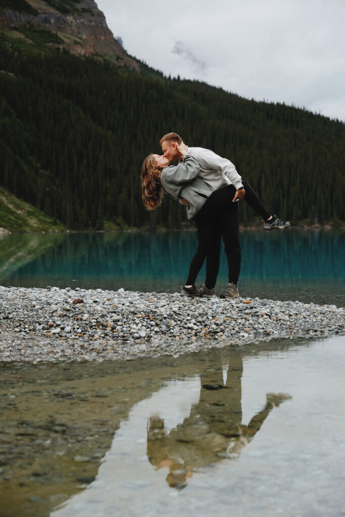 Couple kisses on a shoreline in Alberta. Crystal blue lake in the background with mountains.