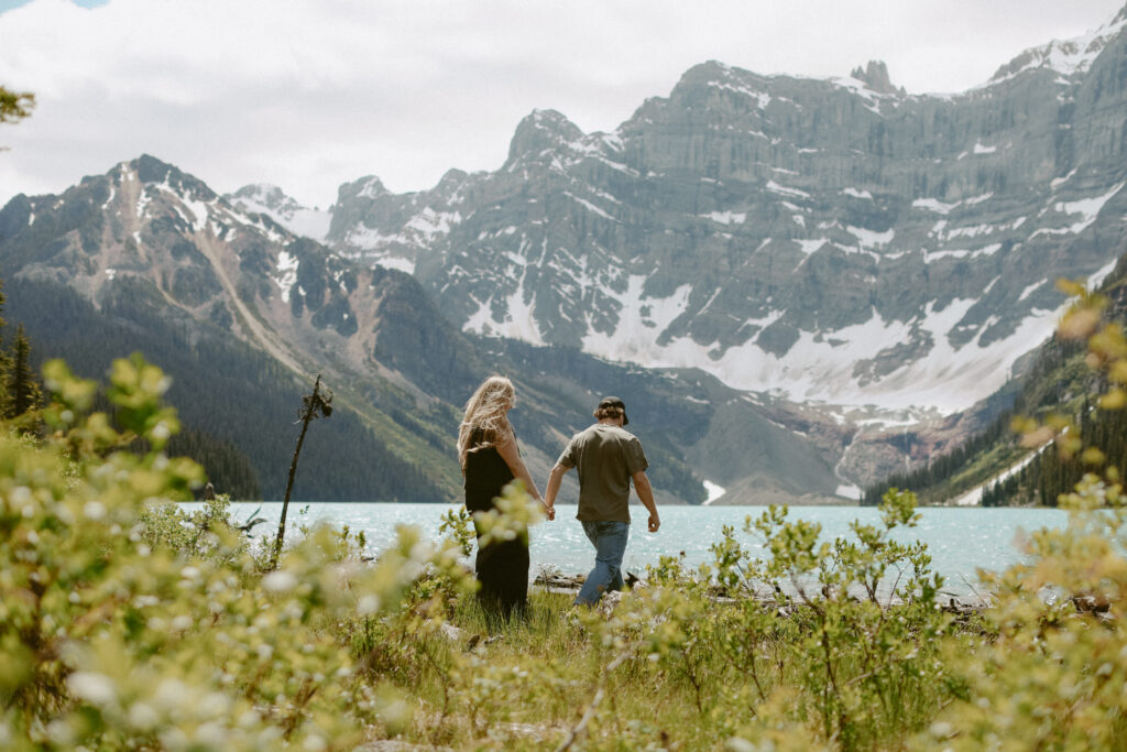 Couple walks hand in hand toward lake with mountains in the background