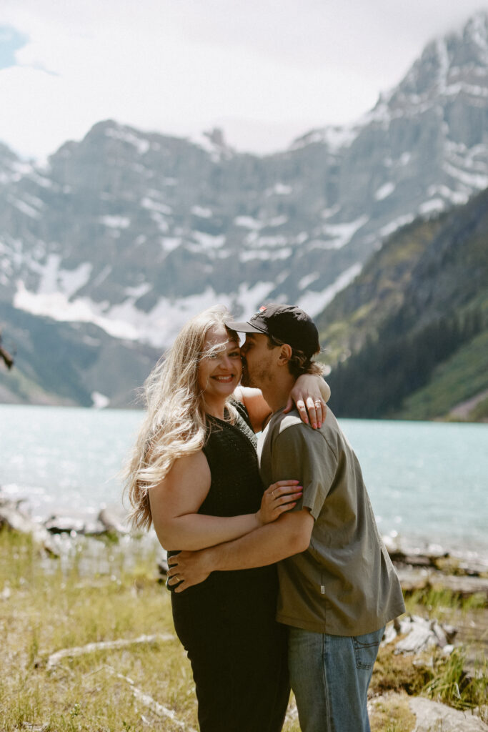 Man kisses women on the cheek during engagement photoshoot with mountains and a lake in the background