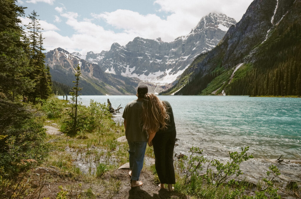 Couple leans on each other during engagement photoshoot with mountains and a lake in the background