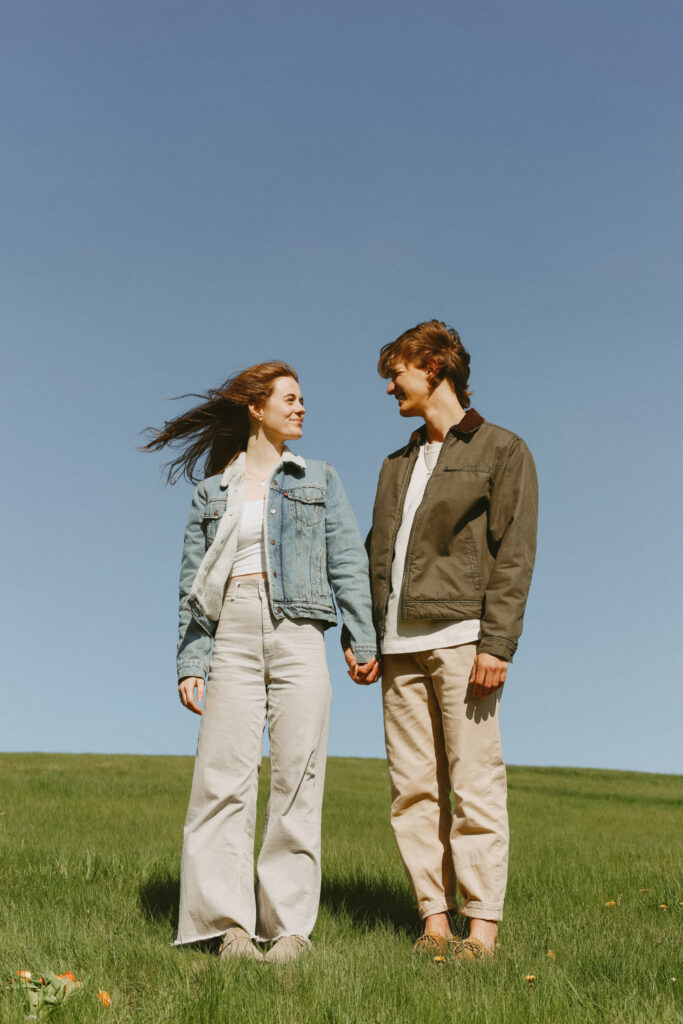 Couple holds hands on a grass hill. The wind blows their hair. 