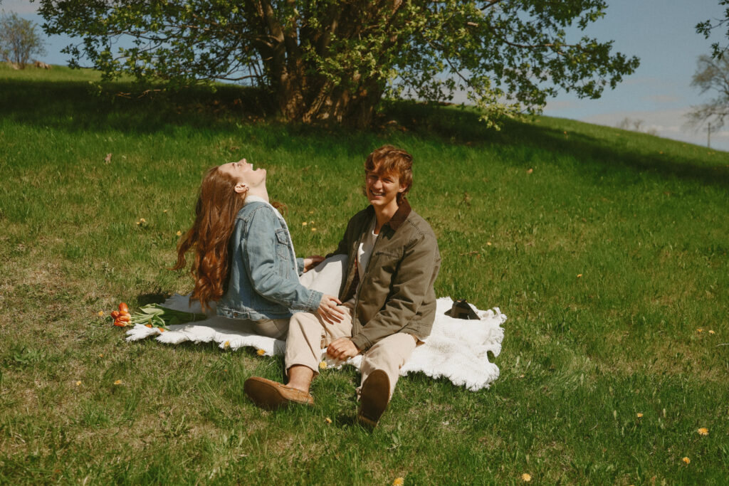 Couple sits on white blanket outside on a grass landscape. 