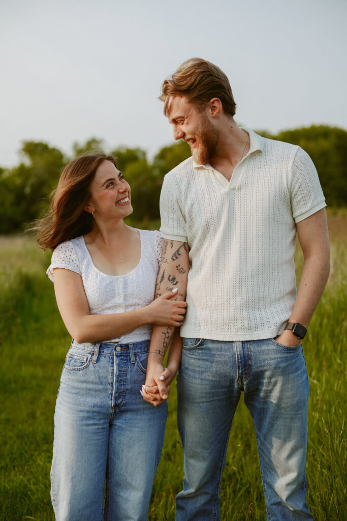 Couple holds hand in field while smiling and looking at each other