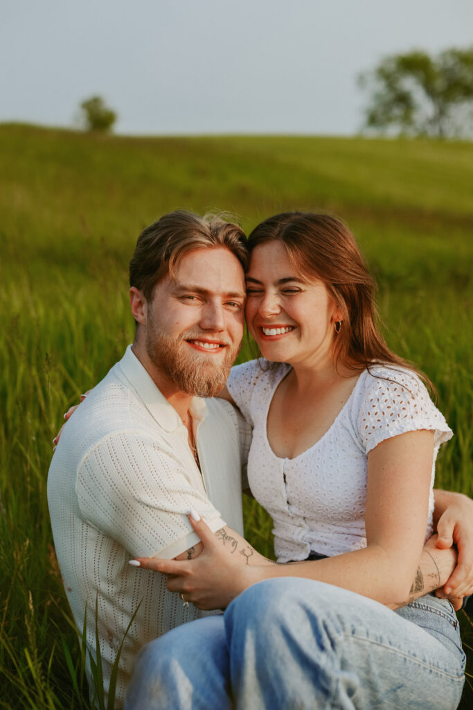 Couple sits closely together in a field. Engagement photography.