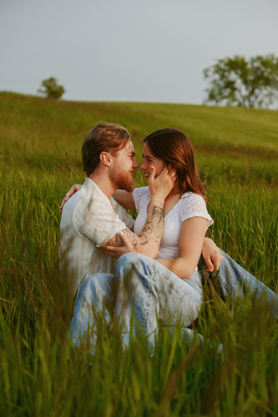 Couple sits closely together in a field. Engagement photography.