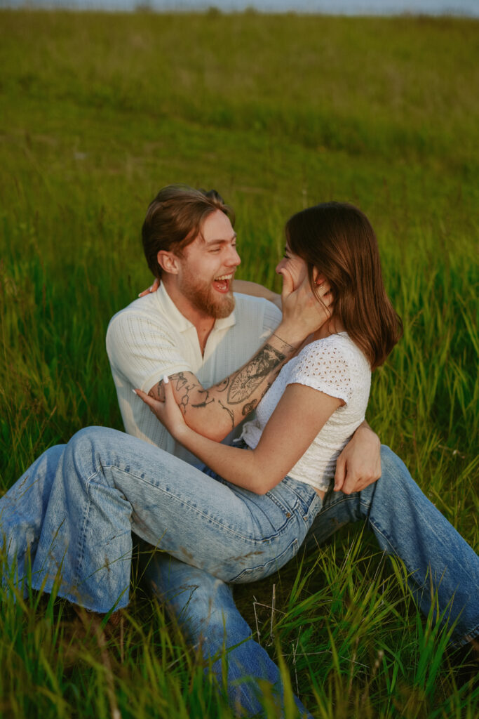 Couple sits closely together in a field. Engagement photography.
